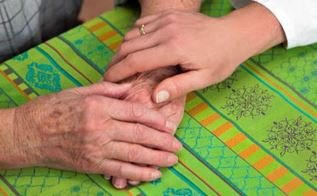 Et par ældre hænder ligger follede på et bord med en grøn dug. Ovenpå hænderne ligger en yngre hånd.
